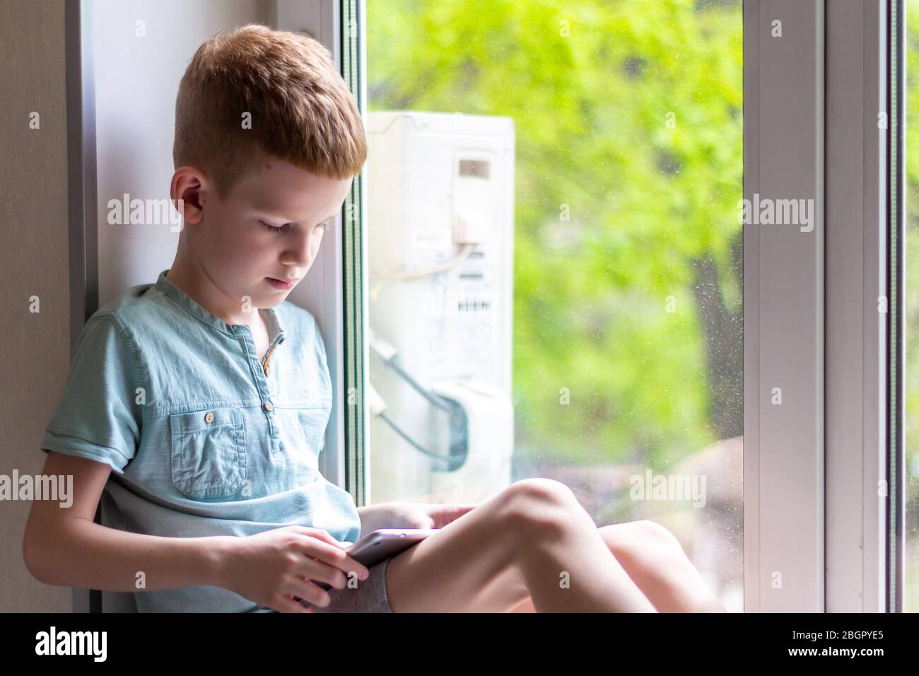 Young boy sitting on windowsill with tablet and play the games. Stay at home during quarantine. Stock Photo