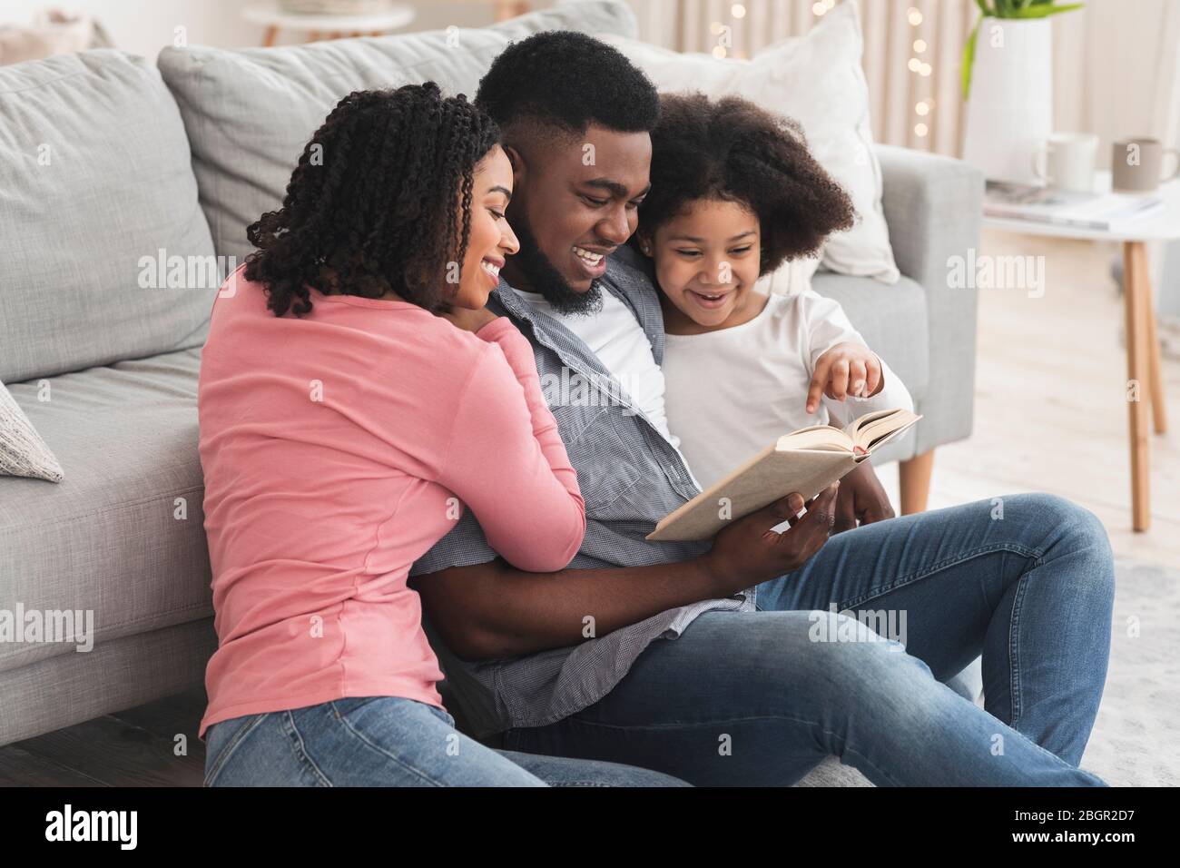 Happy black family reading book together, sitting on floor in living room Stock Photo