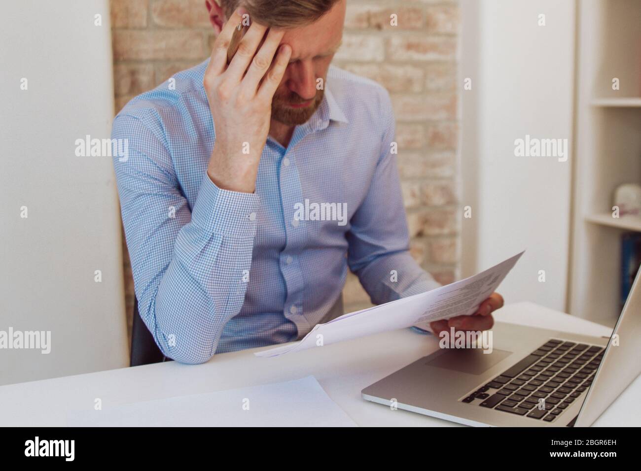 Man sitting at a desk with laptop and reading papers in light blue shirt. Light workspace, business space. Working environment. Brick wall background. Stock Photo