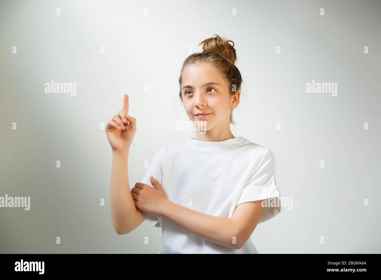 Portrait beautiful young american girl standing finger pointing up something on white gray wall background, child people concept. Natural sunlight. Stock Photo