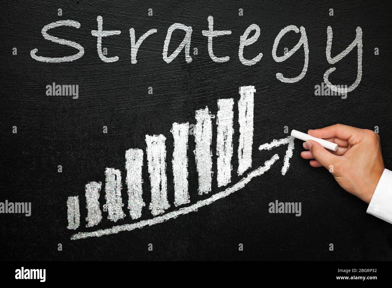 Blackboard with handwritten strategy text. Arrow going upwards and stack chart. Hand with chalk in hand. Direction and success concept. Stock Photo