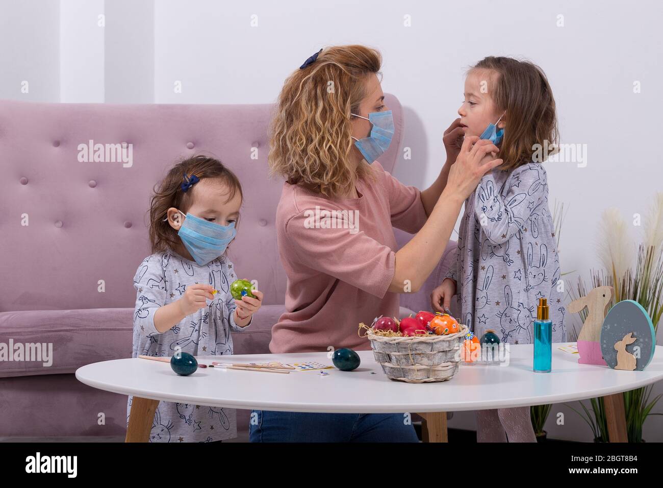 Mother and daughters are painting eggs. Happy family are preparing for Easter. Mother wearing her daughter's mask against coronavir. Quarantine at hom Stock Photo