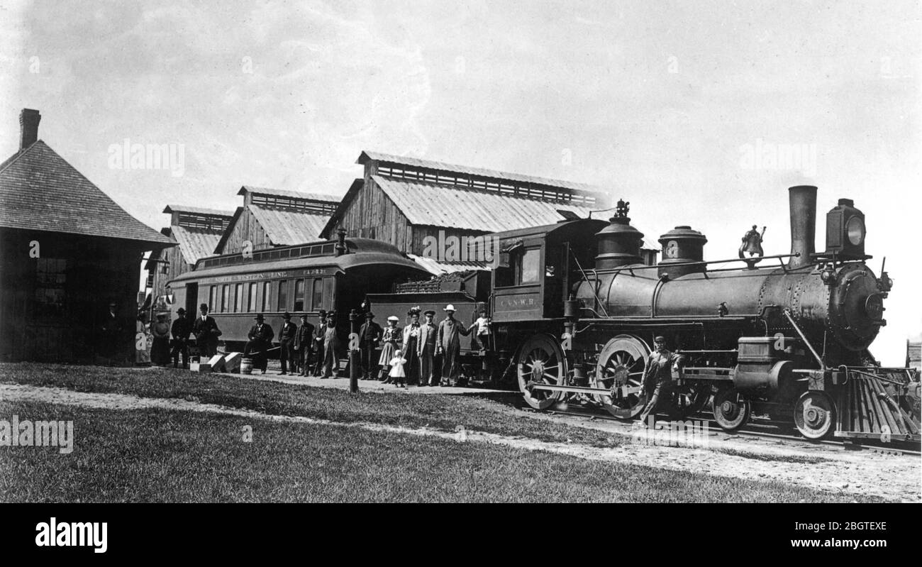 In the 1910s, a steam locomotive, with its coal car and one single passenger car at a small town train depot in northern Wisconsin, America. There is no caboose attached, because this would have been a small train used for short runs between many northern towns and villages. The station depot building, on the left, is dwarfed by three large barns across the tracks. They were probably used for storing corn or farm products that needed drying. Fifteen passengers are waiting, while one engineer stands by the engine. To see my transportation-related images, Search: Prestor  vintage vehicle Stock Photo
