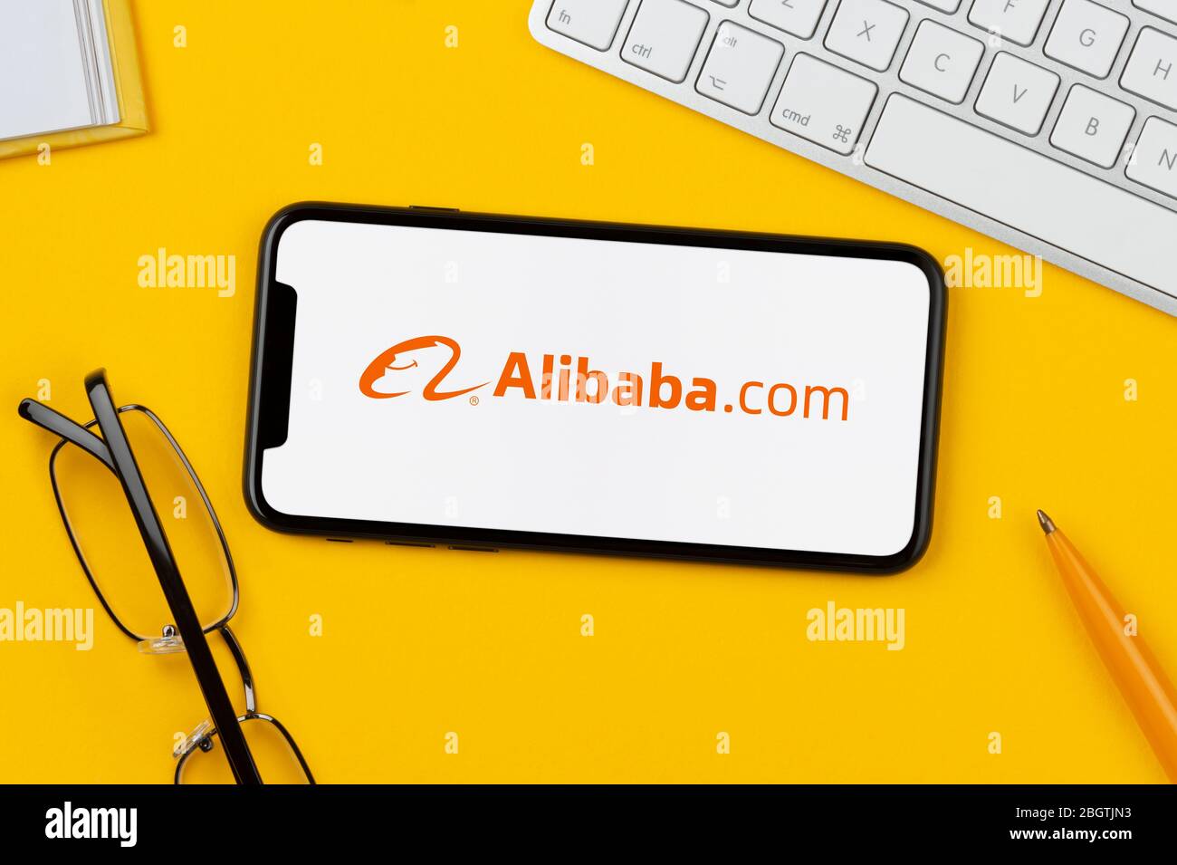 A smartphone showing the Alibaba logo rests on a yellow background along with a keyboard, glasses, pen and book (Editorial use only). Stock Photo