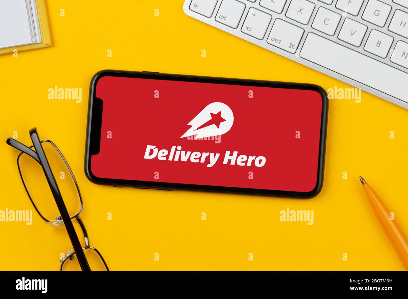 A smartphone showing the Delivery Hero logo rests on a yellow background along with a keyboard, glasses, pen and book (Editorial use only). Stock Photo