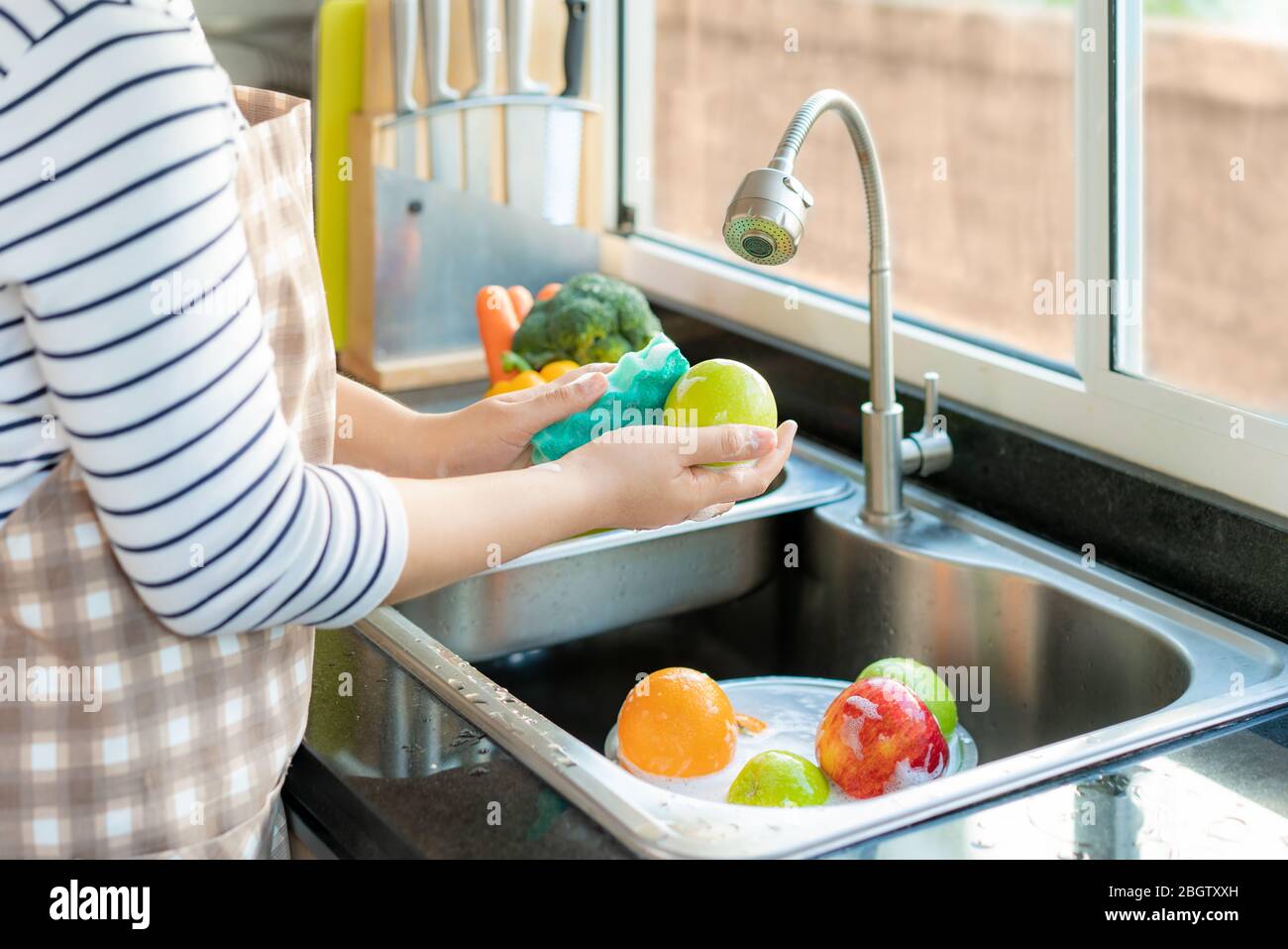Asian healthy woman washing an apple and other fruit above kitchen sink and applying a fruit / vegetable with soap to eliminate the chances of contami Stock Photo