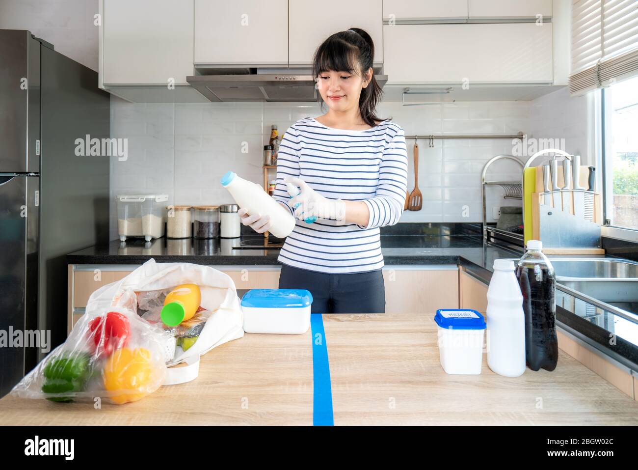 Asian young woman laying out groceries on a divided table and wiping down yogurt or milk containers bottle with disinfectant take to eliminate the cha Stock Photo