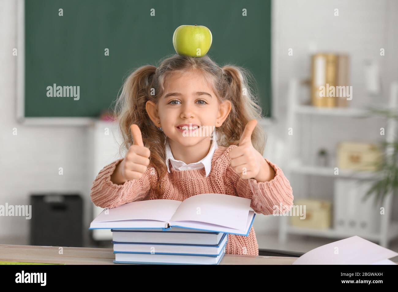 Cute little schoolgirl with books and apple showing thumb-up at desk in ...