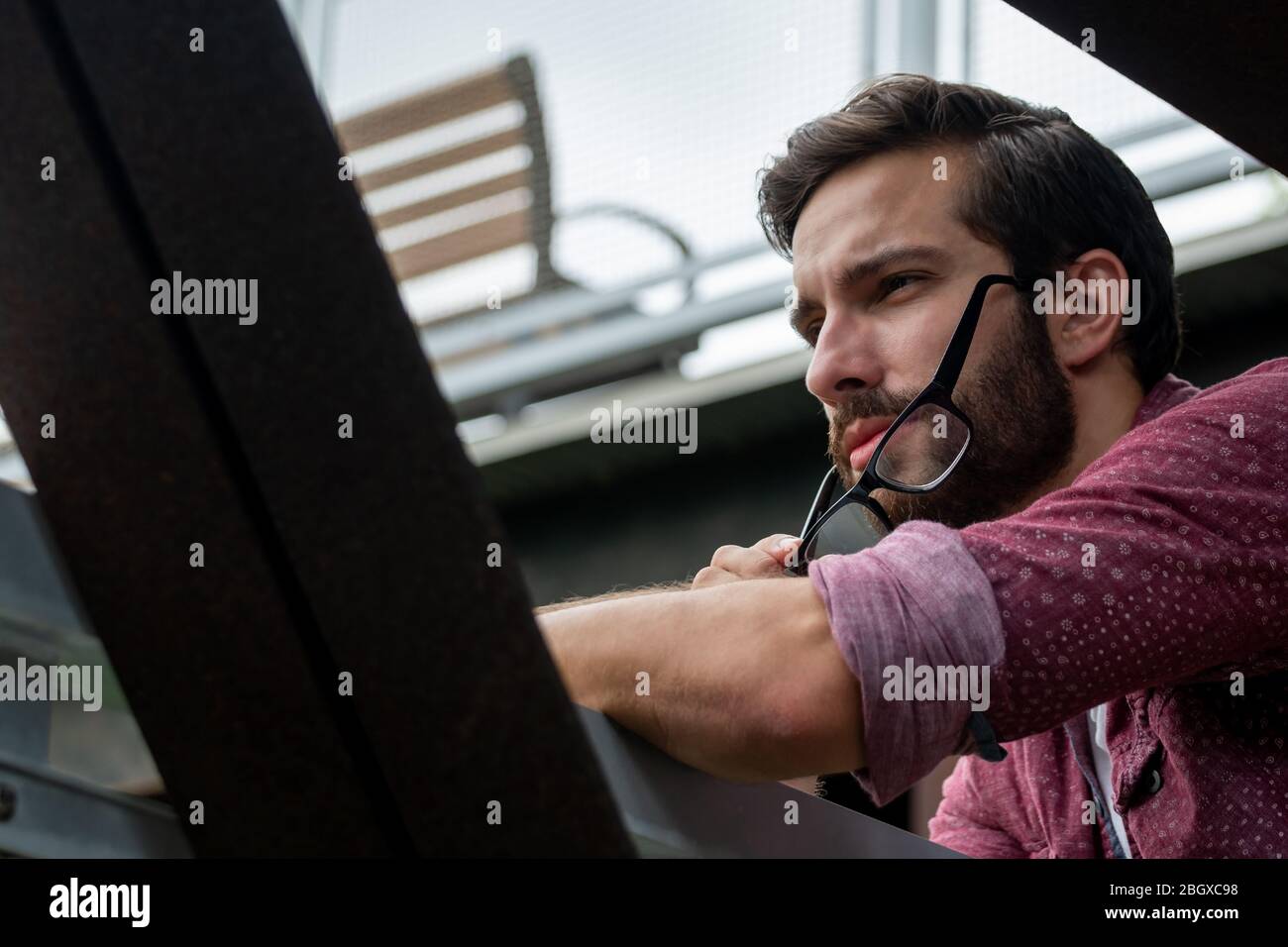 A bearded hipster man wearing a red button down shirt slightly taking off his glasses to see more clearly what is in the distance with a straight star Stock Photo