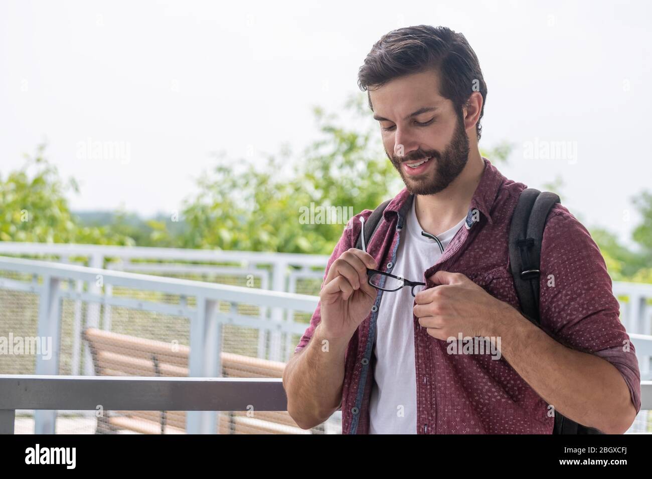 A bearded hipster man wearing a red button down shirt slightly taking off his glasses to see more clearly what is in the distance with a straight star Stock Photo