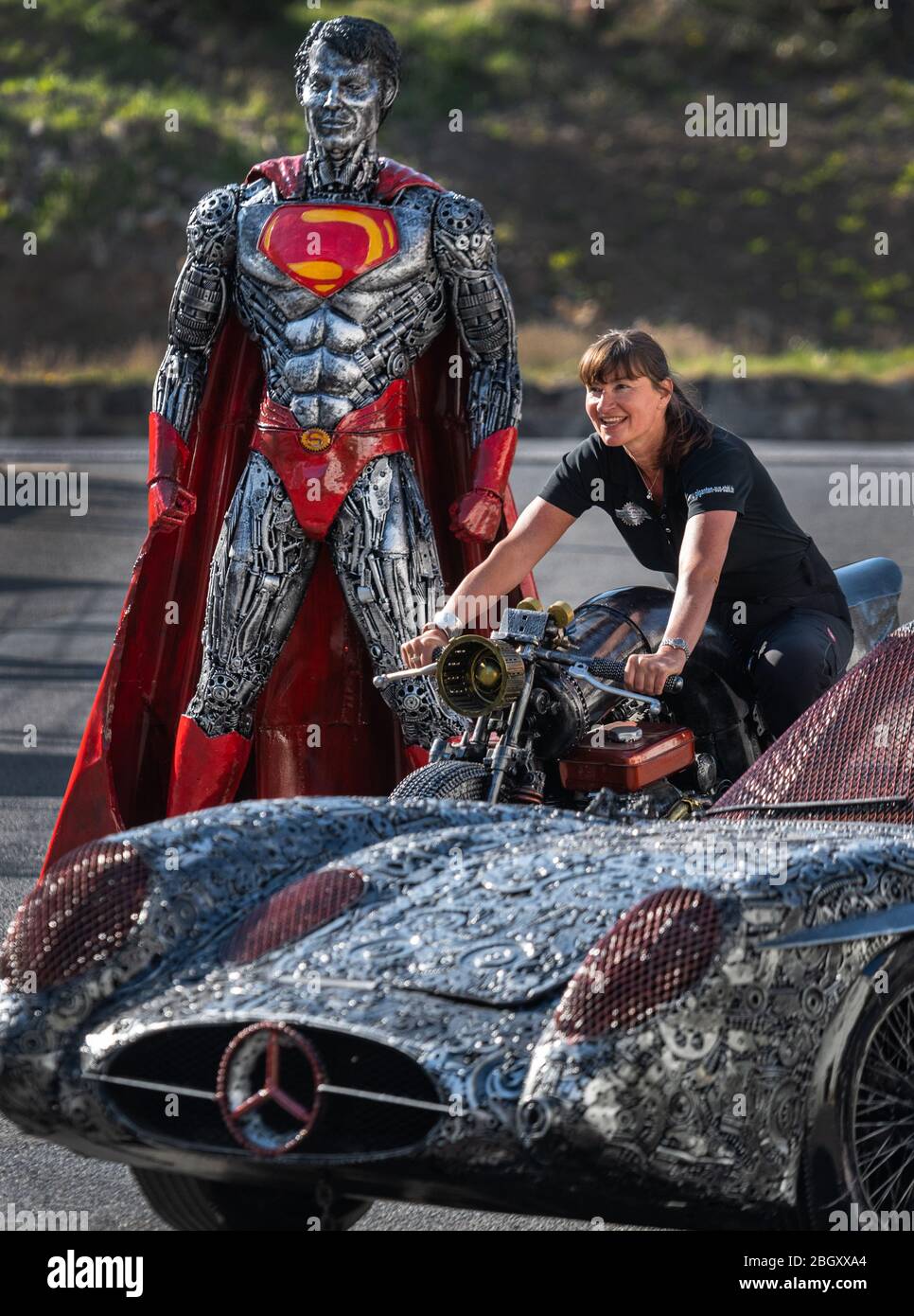 21 April 2020, Saxony, Dorfhain: Steffi Glück, team member of the 'Giants of Steel' is sitting on the grounds of the charitable foundation Georado, on a motorcycle made of scrap metal, behind it stands 'Superman' and in front of it 'Mercedes SLR 300 Uhlenhaut Coupe'. The foundation also uses art to enhance the region. For this purpose, the Geoart network was created, which unites artists with the public and sculptures. For a fair, which cannot take place in its original form due to the Corona crisis, the foundation has brought parts of the 'Giants of Steel', sculptures made from thousands of i Stock Photo