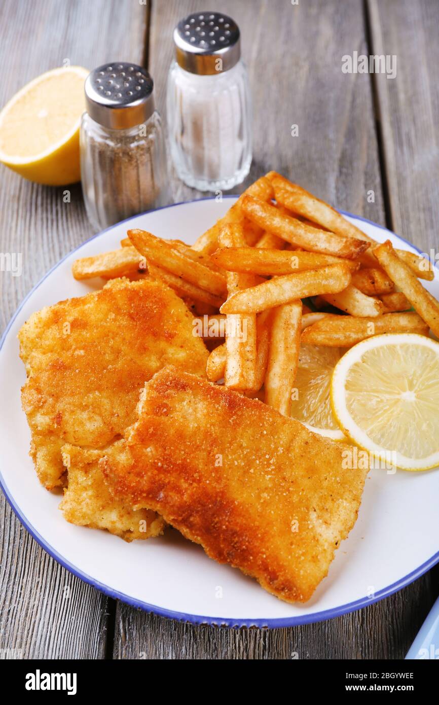 Breaded fried fish fillets and potatoes with with sliced lemon and cutlery on plate and wooden planks background Stock Photo