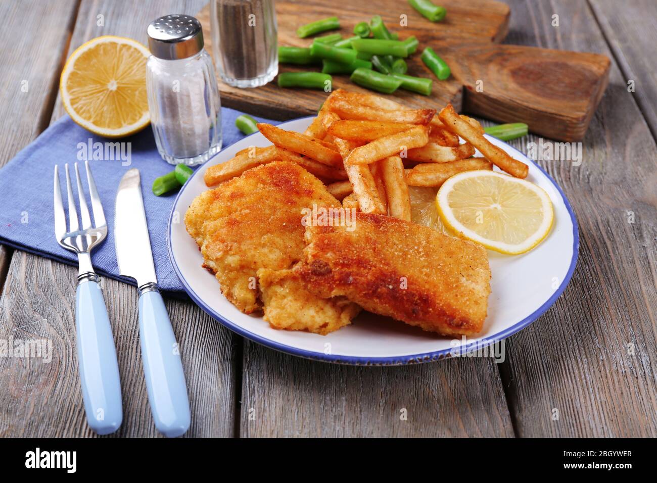 Breaded fried fish fillets and potatoes with asparagus and sliced lemon on plate and wooden planks background Stock Photo