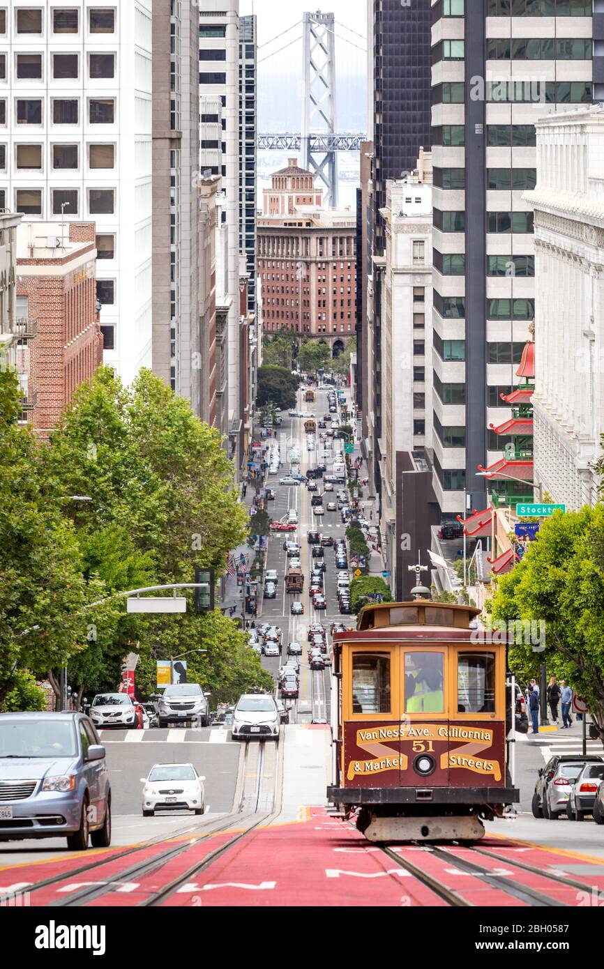 San Francisco, CA - June 16th 2019: a traditional cablecar is running downtown on a sloping street; in the distance, the Bay Bridge Stock Photo