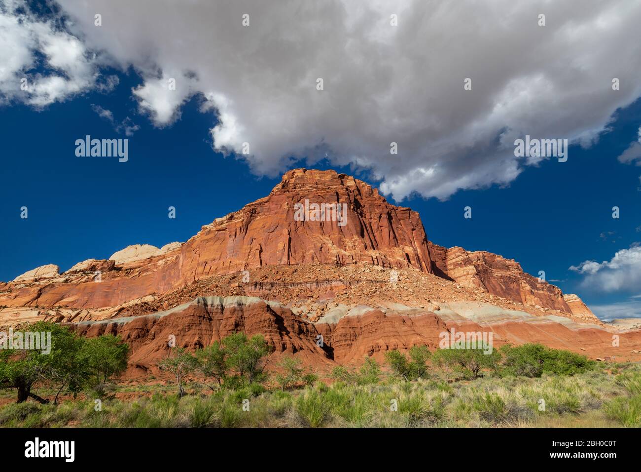 Landscape shot of a large red sandstone formation sloping down on both sides, with a similarly shaped puffy cloud just above it, against a solid blue Stock Photo