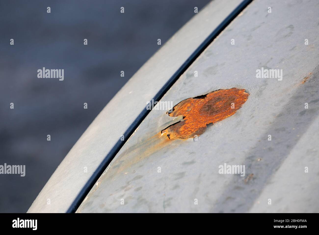 Big orange rust spot on the hood of a silver car. Peeled layer of paint enabling rust and