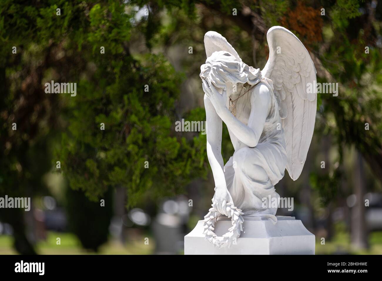Close up of the white marble statue of a kneeling winged angel holding a laurel crown and crying, against a green bokeh background Stock Photo