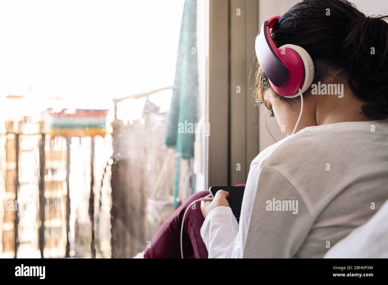little girl sitting in front of her house balcony with headset watching videos on the phone, home entertainment for children concept, copy space for t Stock Photo