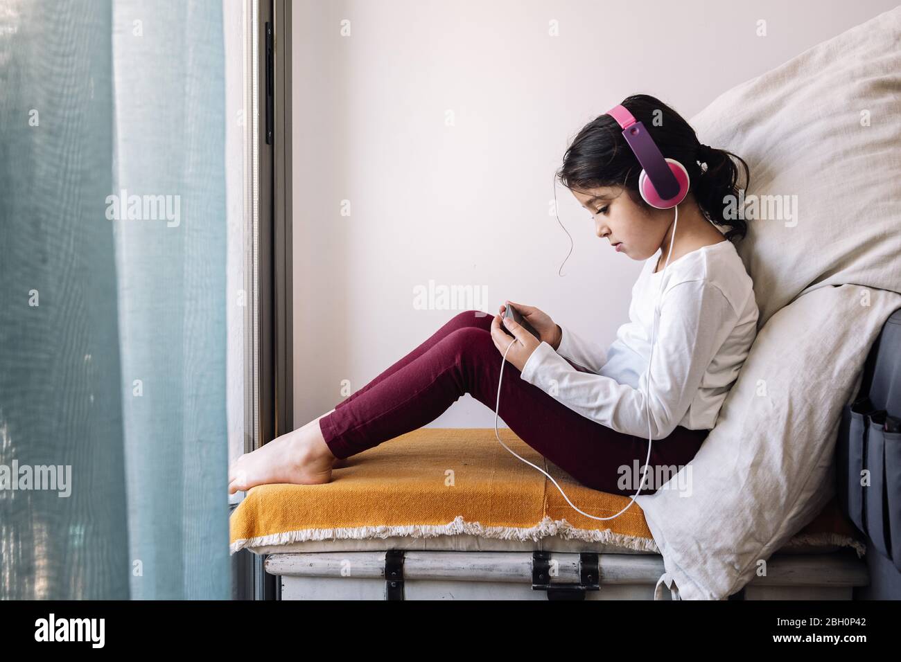 little girl sitting in front of her home window with headphones watching videos on the phone, home entertainment for children concept Stock Photo
