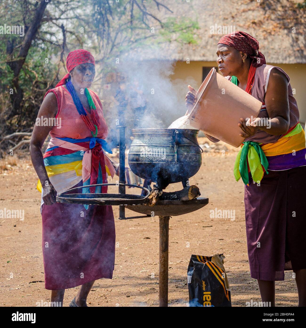African ladies in traditional bright clothing dresses making a soup in a large cauldron over hot charcoal braai Kruger Safari Park South Africa Stock Photo