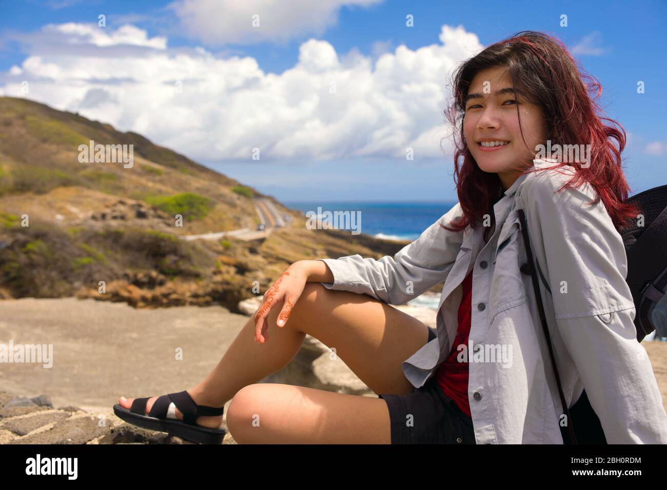 Biracial Asian teen girl sitting on rock wall  looking out over beautiful blue ocean on Oahu, Hawaii Stock Photo