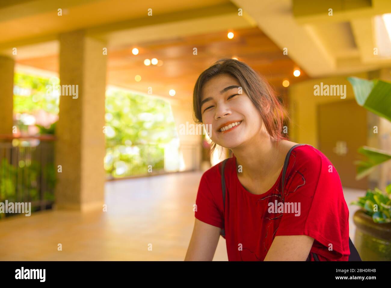 Smiling biracial Asian Caucasian teen girl or young woman sitting in empty outdoor open air lobby in tropical sunny Hawaii, wearing red shirt Stock Photo