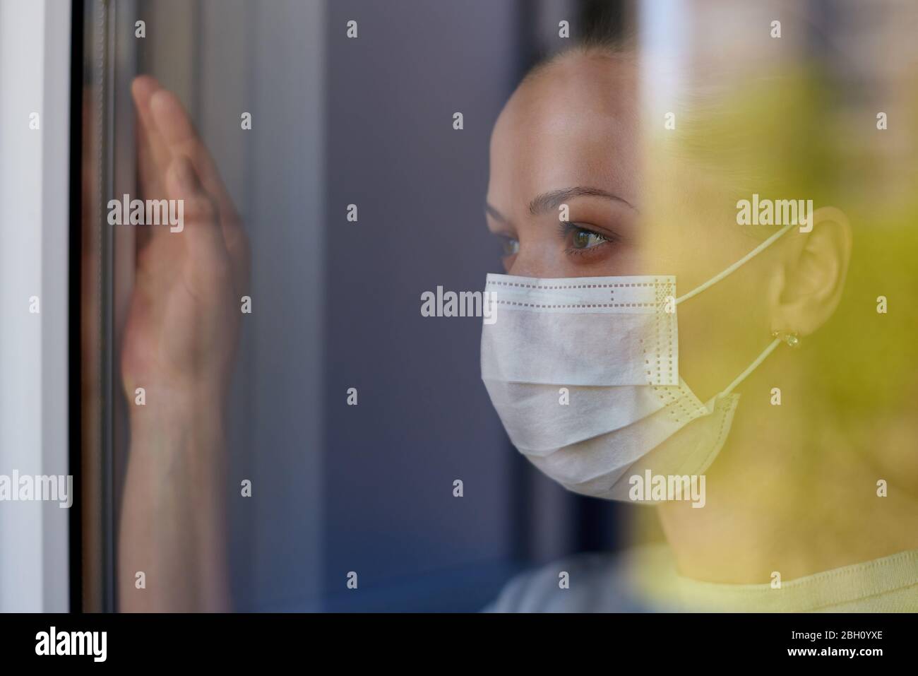 A young sad woman in a medical mask in quarantine looks out the window ...