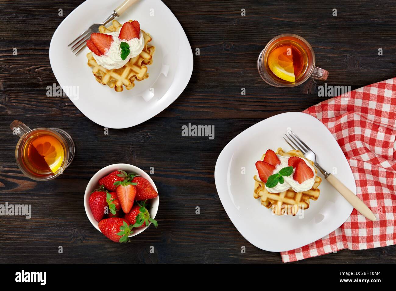 Beautiful breakfast. Belgian Viennese waffles decorated with cream and strawberries, tea and a checkered napkin on a dark wooden rustic background. Stock Photo