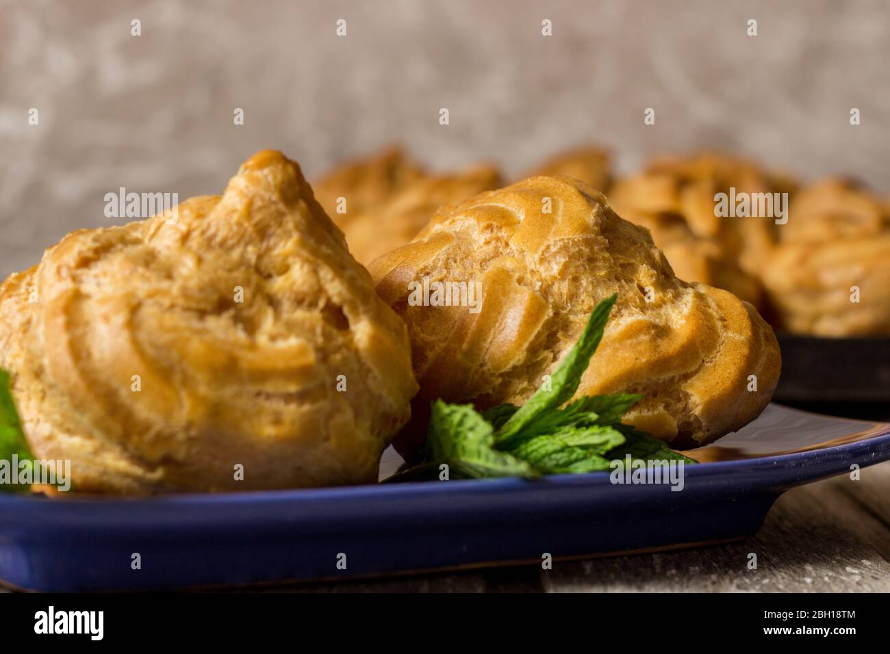 Delicious big cream puffs with cream in hand painted blue oval saucer with mint leaves on aged wooden background. Selective focus. Stock Photo