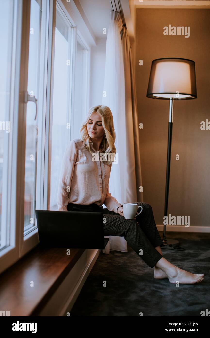 Pretty young woman working on laptop in living room on the window sill ...