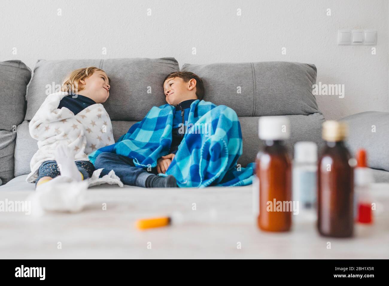 Sick siblings sitting on the couch at home with medications on tabletop