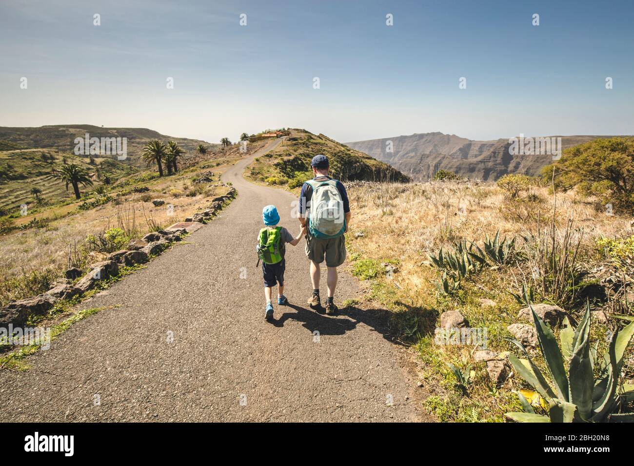 Back view of father and little son with backpacks walking hand in hand on country road, La Gomera, Canary Islands, Spain Stock Photo