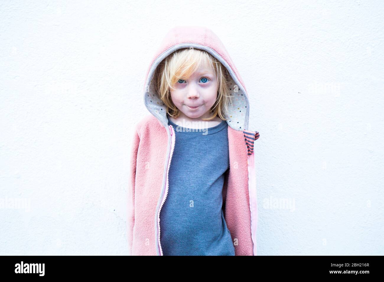 Portrait of little blond girl wearing pink hooded jacket in front of white wall Stock Photo