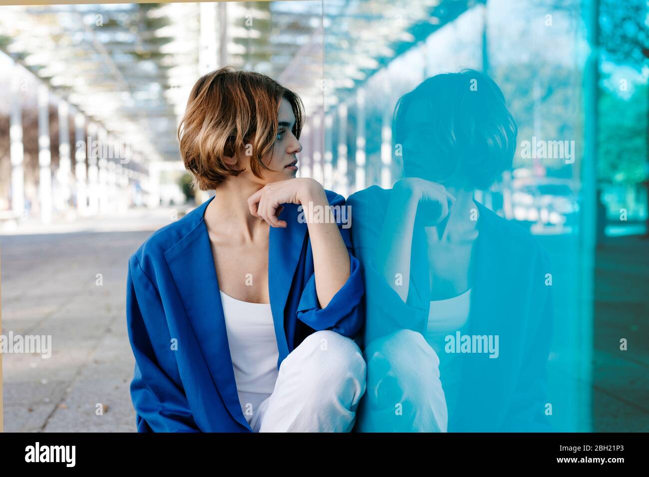 Young woman with urban look sitting on the floor and leaning on colorful glass wall with her reflection Stock Photo