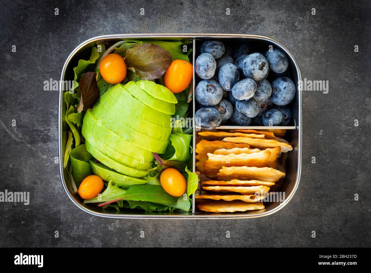 Lunch box with sliced avocado, yellow tomatoes, crackers, blueberries and green salad Stock Photo