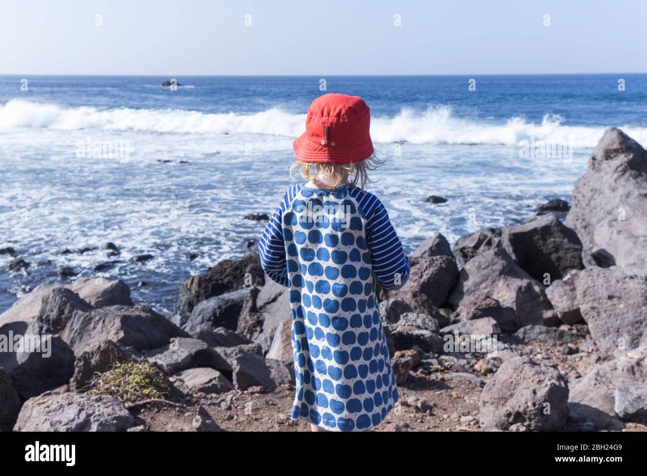 Back view of little girl standing at water's edge looking at the sea, La Gomera, Canary Islands, Spain Stock Photo