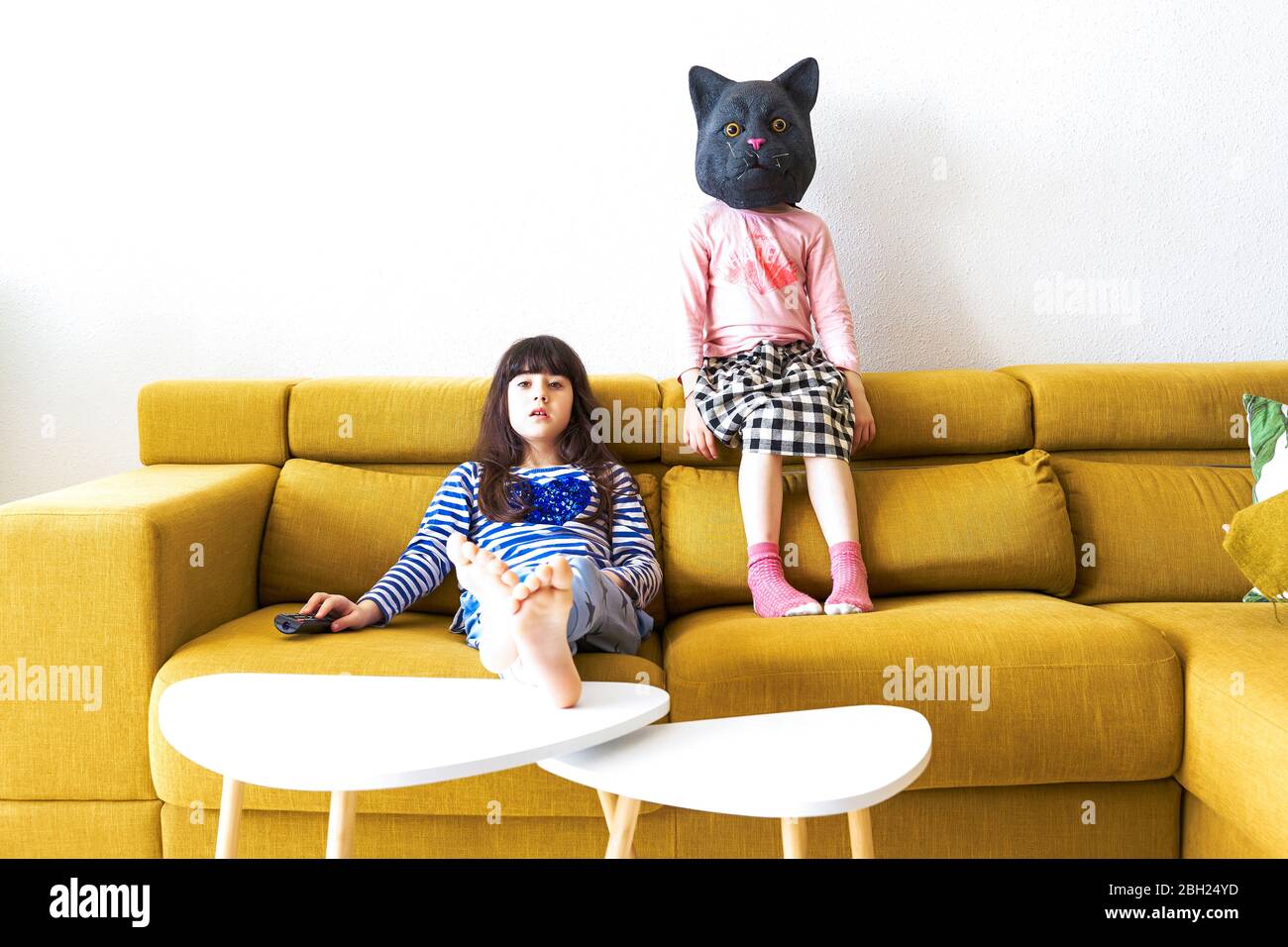 Two bored girls sitting on couch, watching TV, one wearing cat mask Stock Photo