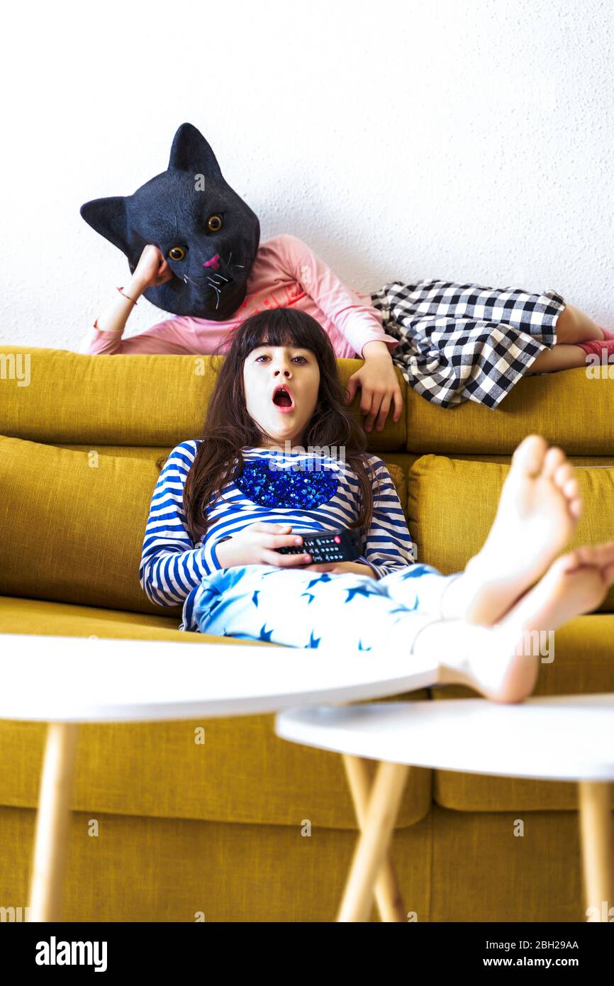 Two bored girls sitting on couch, watching TV, one wearing cat mask Stock Photo
