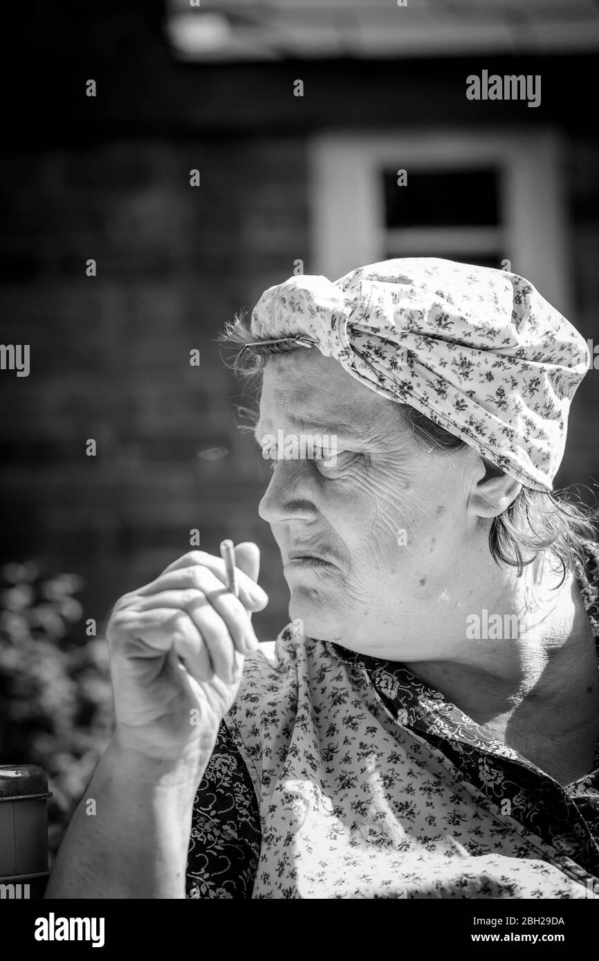 Monochrome, close portrait, 1940s woman as vintage UK housewife, washerwoman, in overalls isolated outdoors smoking cigarette, 1940s WWII summer event. Stock Photo