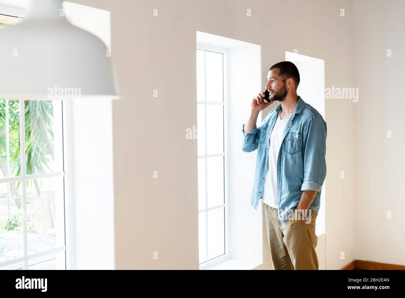 Young man at home in living room speaking on his smartphone while looking out of his window Stock Photo