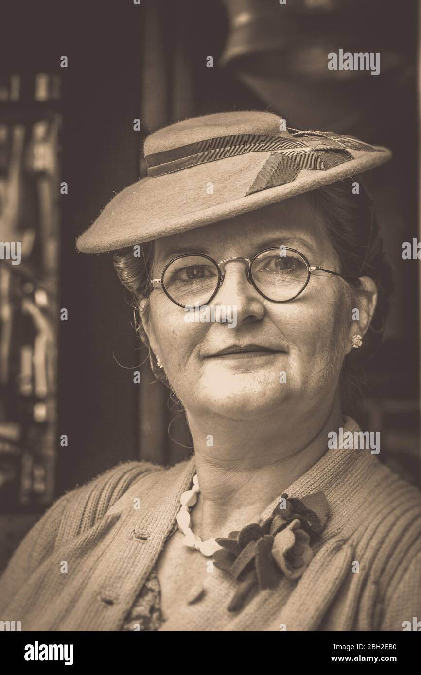 Old-fashioned, vintage sepia front portrait of woman smartly dressed in wartime hat & glasses isolated at UK 1940s WWII reenactment event. Stock Photo
