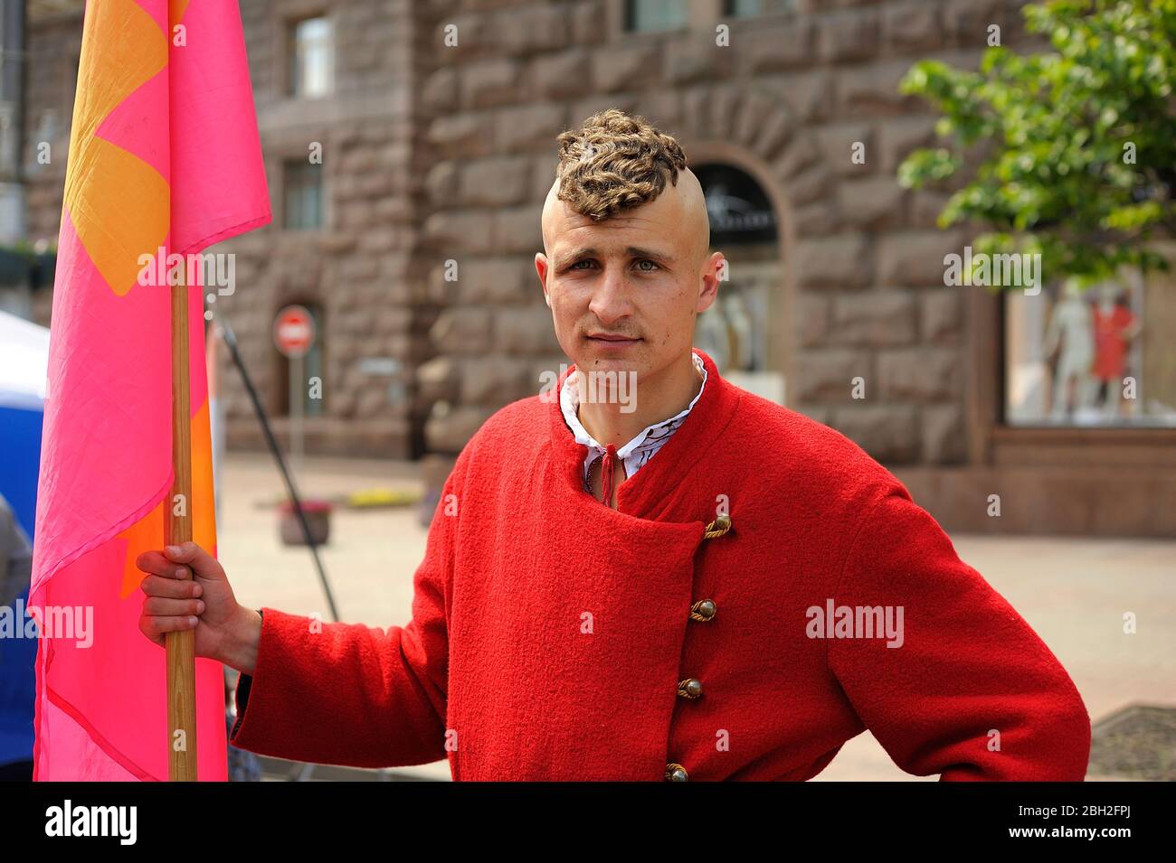 Young handsome man wearing Ukrainian national cossack costume and hairstyle standing on the street and holding flag Stock Photo