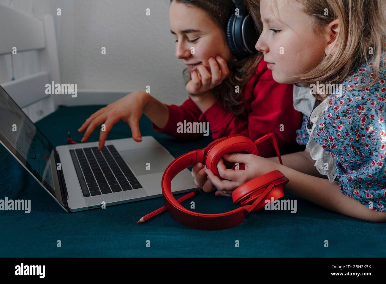 Two sisters with headphones lying on bed using laptop Stock Photo