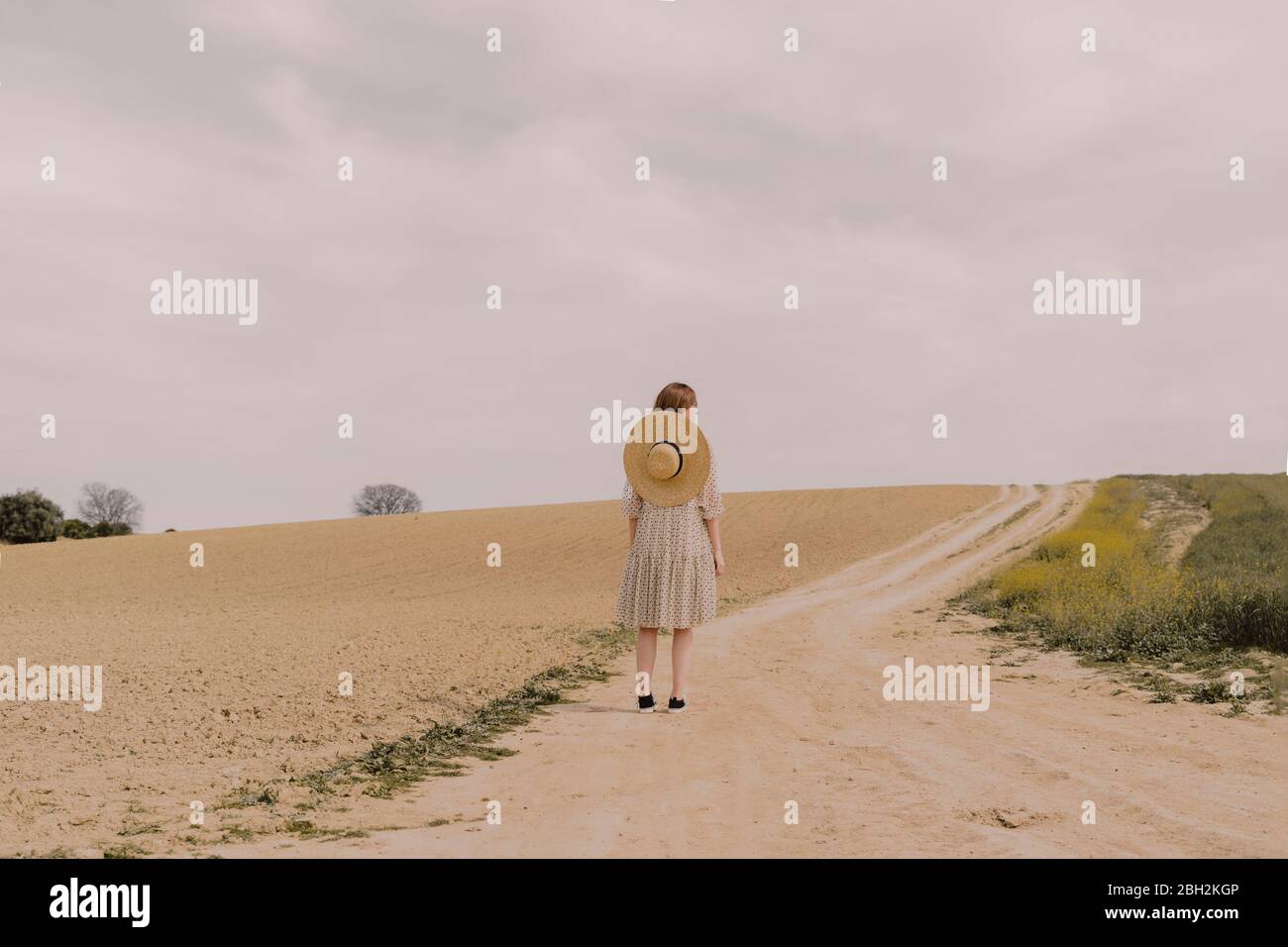 Woman with straw hat and vintage dress alone at a remote field road in the countryside Stock Photo