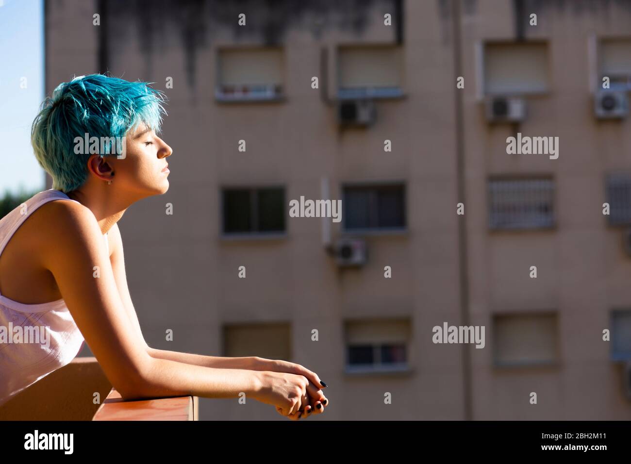 Androgyne young woman with blue hair enjoying sunlight on balcony Stock Photo