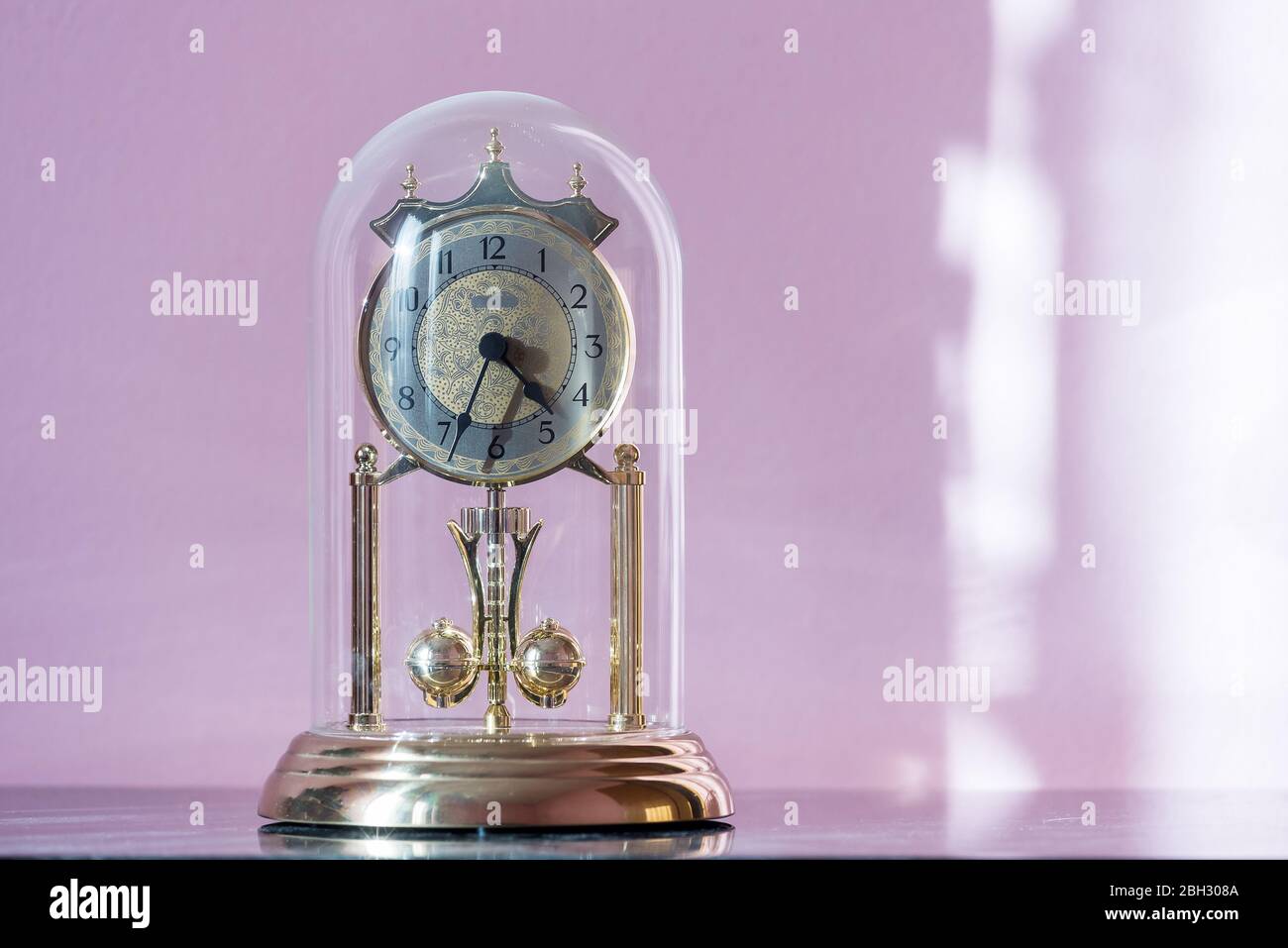 Mantel Clock with Glass Dome & Rotating Pendulum. Period clock with oscillating mechanism on an old dusty Cupboard lit by morning sun Stock Photo