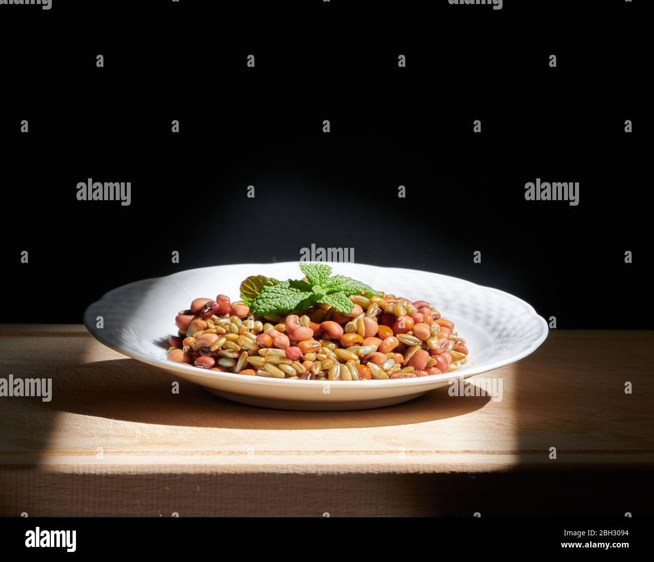 Beans and rye with mint leaves on a white plate, on a wooden chopping board, illuminated by direct sunlight Stock Photo