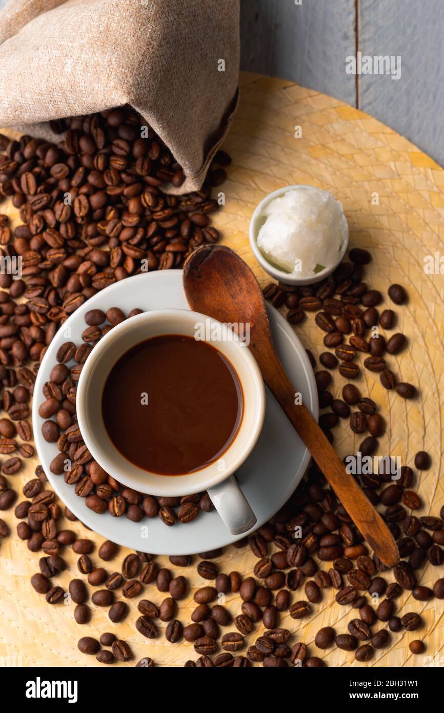 White cup and saucer with coffee and surrounded by roasted coffee beans on a wooden table and a bag next to a small cup of coconut oil and a wooden sp Stock Photo