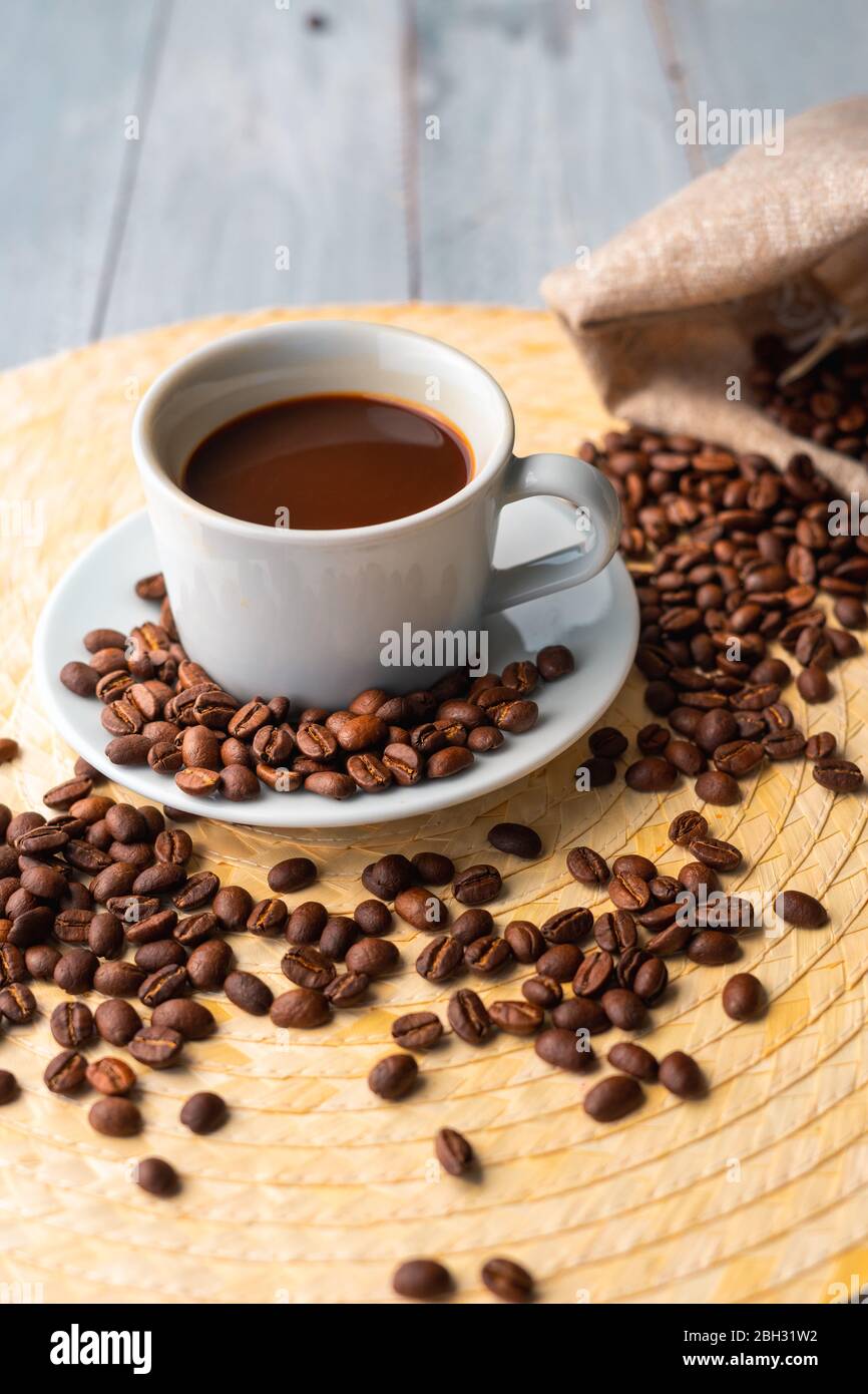White cup and saucer with coffee and surrounded by roasted coffee beans on a wooden table and a bag Stock Photo