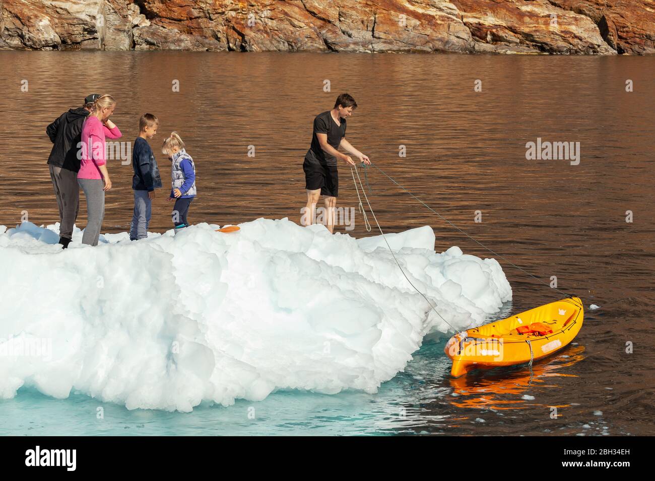 A mixed group, including a child, standing on an iceberg floating in sea, with a yellow kayak next to the iceberg Stock Photo